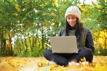 Girl in hipster with laptop in autumn park. A woman in a cap using a laptop while sitting on fallen leaves. Freelancer in the hat uses remote communication technology. Remote work. Free space
