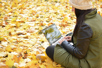 Girl in hipster with laptop in autumn park. A woman in a cap using a laptop while sitting on fallen leaves. Freelancer in the hat uses remote communication technology. Remote work. Free space