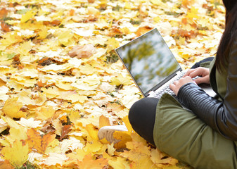 Girl in hipster with laptop in autumn park. A woman in a cap using a laptop while sitting on fallen leaves. Freelancer in the hat uses remote communication technology. Remote work. Free space