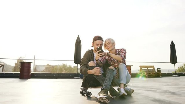 Handsome young hipster couple are sitting on longboard skateboard on the rooftop of modern building and using smartphone. Two young people making selfie photo on smartphone. Concept of modern urban