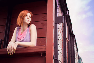 The red-haired girl with a short haircut is standing on an old, wooden train. Old school train.