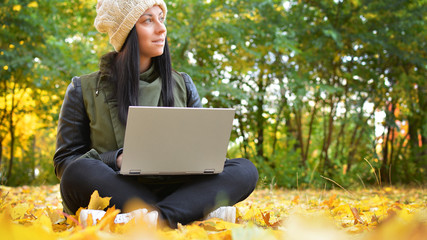 Girl in hipster with laptop in autumn park. A woman in a cap using a laptop while sitting on fallen leaves. Freelancer in the hat uses remote communication technology. Remote work. Free space