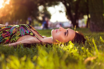 Gorgeous young beautiful girl lies on the grass in the park. Woman with beautiful make-up in a colored dress resting in a park. Dream.