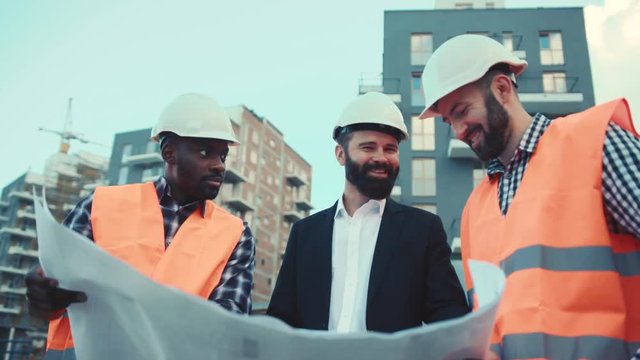 Cheerful multiracial team of construction managers and handsome bearded businessman in hard hats compare the project with the work done. Teamwork, cooperation.