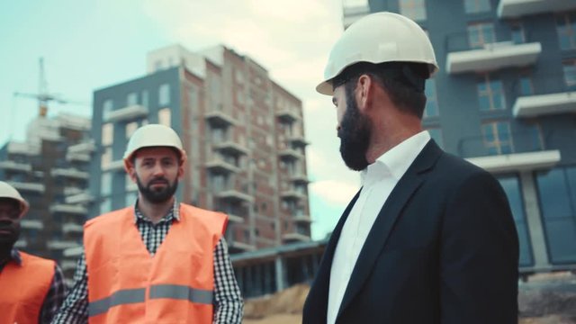 Multiracial professional construction workers having a meeting with a young serious businessman in luxurious suit, discussing a project. Making money, cooperation.