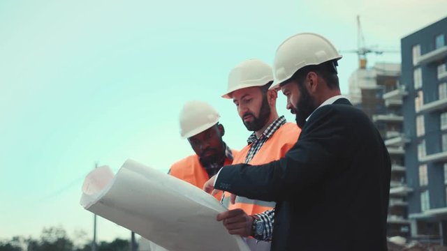 Low angle view of handsome bearded businessman and multiracial builders with blueprint looking away, discussing project. Team work, profession concept.