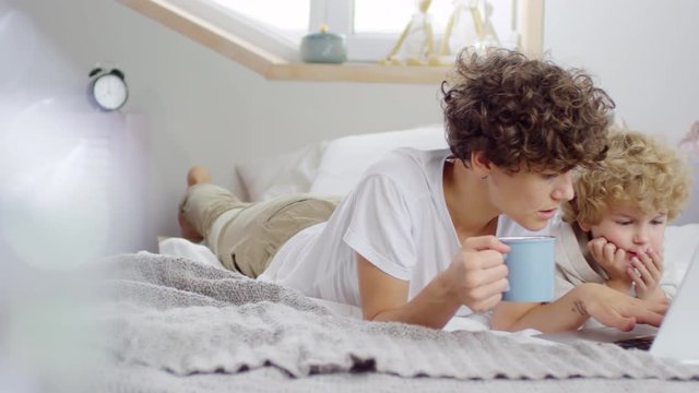 PAN shot of happy mother and little boy with curly hair lying on bed and chatting while looking at laptop