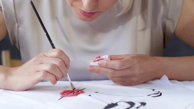 Tilt down shot of creative woman with short blond hair creating fashion illustration on white t-shirt lying on table
