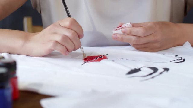 Mid-section panning shot of hands of unrecognizable woman painting fashion illustration of abstract female face with red lips on white t-shirt