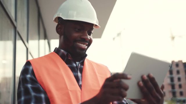 Young handsome smiling Afro-American construction worker in signal jacket and white helmet actively using tablet by the unfinished building. Browsing the internet, profession concept. Male portrait
