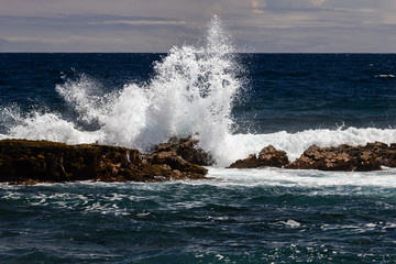 Wave crashing on dark volcanic rock at Black Sand Beach (Punaluuu), on the Big Island of Hawaii. White spray rises in the air; Blue pacific ocean, sky and clouds are in the background. 