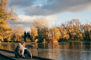 Mother and son sitting near lake in autumn park on sunset and looking away. Family having fun, hugging, laughing, relaxing, enjoy life.Close up sunny lifestyle fashion portrait of happy stylish family