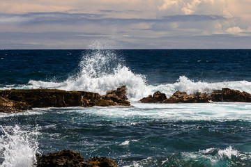 Wave crashing on dark volcanic rock at Black Sand Beach (Punaluuu), on the Big Island of Hawaii. White spray rises in the air; Blue pacific ocean, sky and clouds are in the background. 