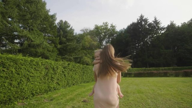 Slow motion of baby girl with mother running away on green grass in the summer light in park. Baby have fun.  Baby laughing. Green thuja background. Happy family concept. Woman with long hair.