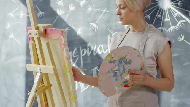 Tilt up shot of female artist with short blond hair painting on easel in studio with chalkboard art on wall