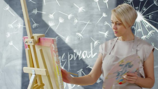 Medium shot of creative woman with short blond hair painting on easel in studio with chalkboard mural on wall