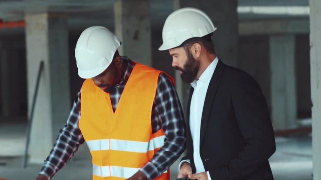 Professional engineer and construction worker checking the blueprint with the real life building outdoors, making changes, comparing with the project on phone. Modern technologies.