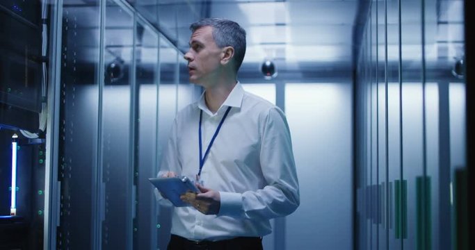Adult IT worker in white shirt standing among server racks in data center smiling at camera