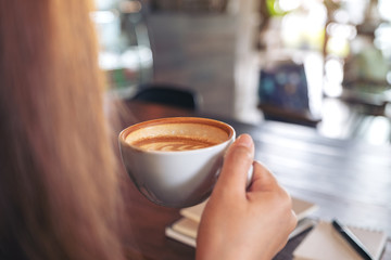 Closeup image of a woman holding and drinking hot coffee with notebooks on table in cafe