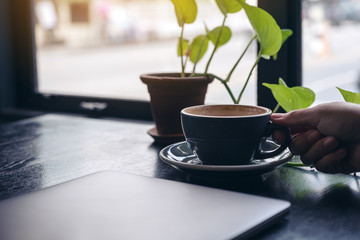 Closeup image of a woman's hand holding a cup of coffee with computer laptop and small tree pot on wooden table