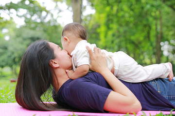 Happy Asian mom embrace her son lying in the green garden. Funny mother and infant baby boy playing in summer park.