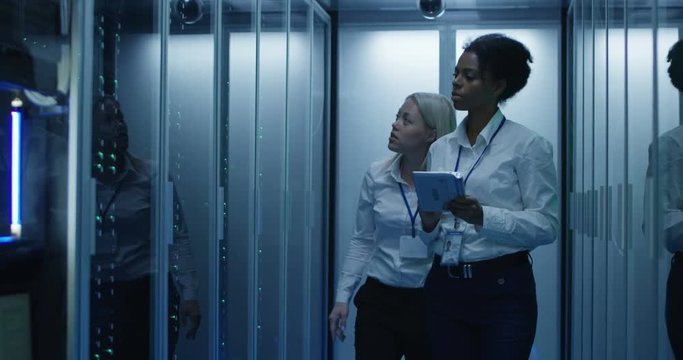 Medium shot of two women working in a data center with rows of server racks and checking the equipment and discussing their work