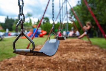 Closeup picture of a swing in a park for kids. Kids swigning in the blurry background - Picture taken on a warm summer day, with mulch ground instead of sand, and green lawn in the background - obrazy, fototapety, plakaty