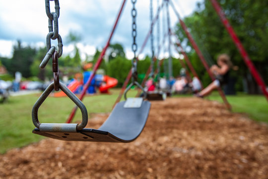 Closeup picture of a swing in a park for kids. Kids swigning in the blurry background - Picture taken on a warm summer day, with mulch ground instead of sand, and green lawn in the background