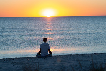 Naklejka na ściany i meble Man sits on the beach next to surf line, practicing yoga