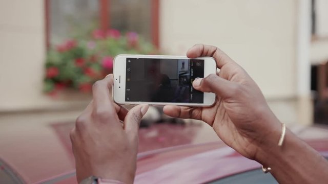 Hands of young African man holding smartphone. Guy taking pictures of cute cat laying on automobile. Parts of body. Summertime. Outdoors.