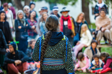 Woman with platted hair and glasses is giving a public speech or conference at the park in front of a blurry crowd of 50 people - 1/5