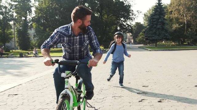 Happy good-looking Caucasian man with beard riding a bicycle in park. Nice son wearing helmet running after daddy. Game in park. Summertime. Outdoors.
