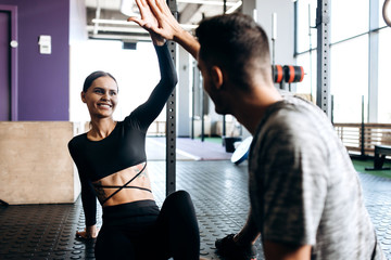 Young athletic man and slim girl are sitting on the floor and give five in the gym