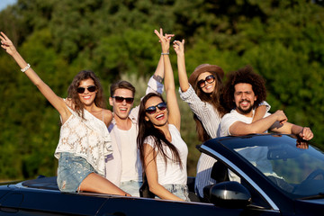 Happy beautiful young  dark-haired girls and guys in sunglasses smile in a black cabriolet on the road holding their hands up on a sunny day.