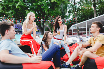 A company of good-looking friends laughing, drinking yellow cocktails and socialising while sitting on the bench and on the lawn in the nice summer cafe. Cheers. 