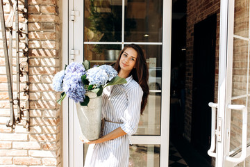 Beautiful brunette girl dressed in a striped dress smiles and and hold a vase with light blue...