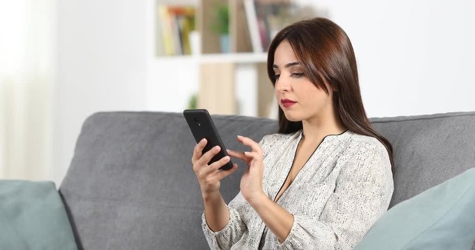 Serious woman browsing content in a smart phone sitting on a couch in the living room at home