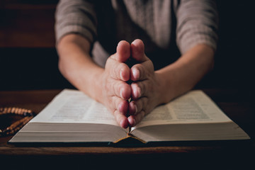 Christian woman praying in church. Hands crossed and Holy Bible on wooden desk.