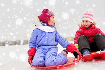 childhood, sledging and season concept - happy little girls on sleds outdoors in winter