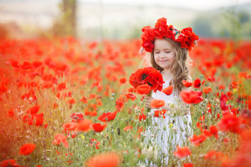 A preschool girl with long curly hair and brown eyes, dressed in a white,spends her time alone in the countryside on a large field with red blooming poppies, wearing a wreath of red flowers