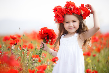 A preschool girl with long curly hair and brown eyes, dressed in a white,spends her time alone in the countryside on a large field with red blooming poppies, wearing a wreath of red flowers