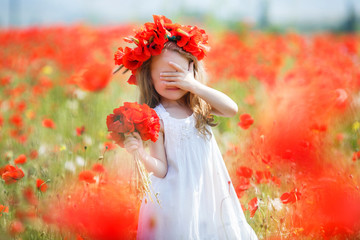 A preschool girl with long curly hair and brown eyes, dressed in a white,spends her time alone in the countryside on a large field with red blooming poppies, wearing a wreath of red flowers