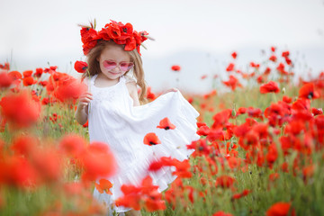 A preschool girl with long curly hair and brown eyes, dressed in a white,spends her time alone in the countryside on a large field with red blooming poppies, wearing a wreath of red flowers