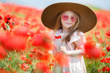 Cute little girl with long curly hair,dressed in a white dress,wears on her head a large straw sun hat,spends time alone in a rural area on a large field with blooming red poppies, holds red flower