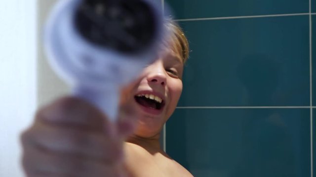 Cute funny little kid having fun in bathroom playing hair dryer. Boy pretending to be shooting from gun into camera, smiling and laughing face. Real time 4k video footage.
