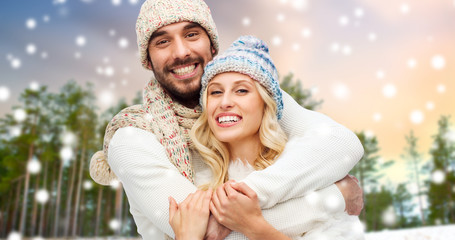 winter, christmas and people concept - happy couple in hats and scarf hugging over forest and snow background