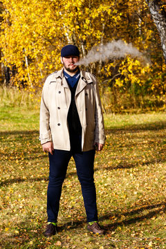 Portrait of handsome casual stylish young man wears trench coat and flat cap smoking vape in colorful autumn forest