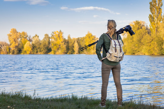 Woman holding a tripod with camera and looking for composition in autumn nature near to lake