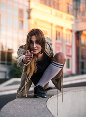 Fototapeta premium Outdoor lifestyle image of a young pretty chestnut haired caucasian girl pointing, sitting, having fun and dancing on the street. Cute casual outfit in front of glass building.