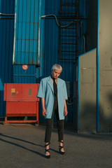 Stylish young eccentric woman with white short hair, wearing blue jacket and black tie, looking down, outside, near colorful metal wall